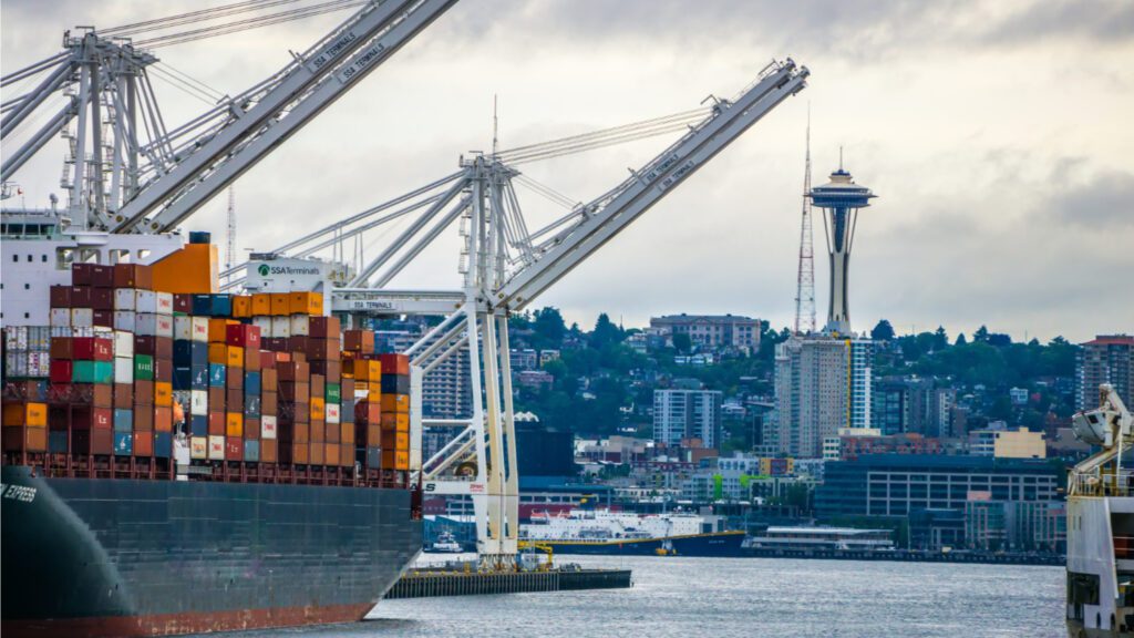 large container ship in foreground on the left side with the Space Needle in the background toward the right and homes in Seattle also in the background